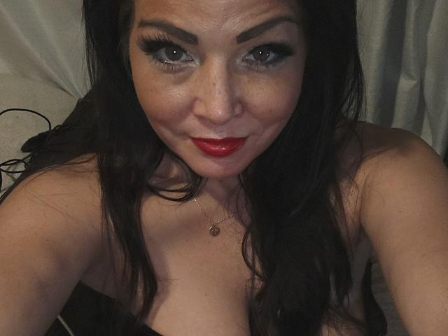 Paiday has dark hair, red lipstick, and thick black eyeliner. She's smiling with a nose ring, wearing a black strap dress, and has a necklace. Boobs are visible.