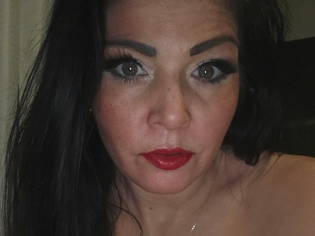 Paiday has dark eyes, black eyeliner, red lipstick, and black hair, with a serious expression, showing her bare shoulders. Her skin is fair, and she's wearing a necklace.