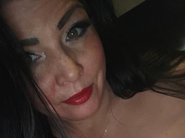 Paiday with dark hair, red lipstick, and heavy eye makeup, has a slightly puffy face and a subtle smile. Her tits are not visible. Her ass is not visible.