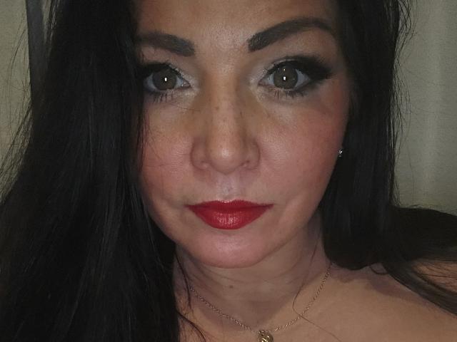 Paiday with dark brown eyes, red lipstick, black hair, and a subtle nose ring, wearing a thin gold necklace, looking directly at the camera with a slight smile.