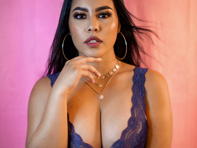 PaolaYKatty with dark hair, wearing a lacy purple top, gold hoop earrings, and a necklace, touching her chin, showing her shiny, tanned, large tits against a pink background.