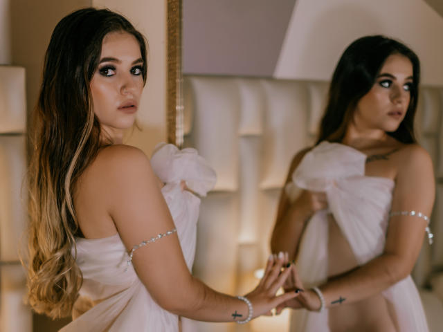 PaolaYKatty con cabello largo y degradado, vestidas con un vestido blanco sin tirantes y con una flor, mirando su reflejo, tomadas de la mano, usando una pulsera de plata, con tatuajes de cruces..