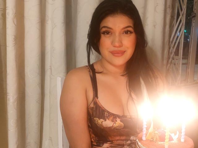 LauraRichis with dark hair, wearing a floral tank top, smiling at a camera with a lit birthday cake in front of her. Her tits are visible, and she's cute.