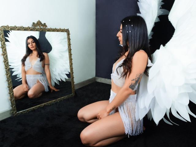 LunaCollinss with white angel wings, kneeling in front of a mirror, wearing a white lace bra and skirt, showing her tits and ass, tattoo on right arm.