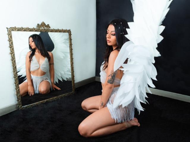 LunaCollinss, kneeling on black carpet, wearing white angel wings and lace lingerie, reflecting in ornate mirror, showing her ass and tattoos, with black and white walls.
