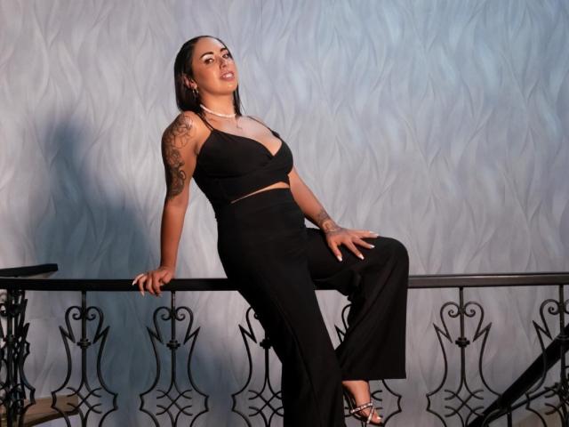 Genexis trägt ein schwarzes Outfit, hat Tattoos am rechten Arm, schwarze High Heels, lehnt sich an ein verziertes Geländer, blickt selbstbewusst nach oben, hat einen hellen Hautton, schwarzes, glattes Haar und wirkt attraktiv..
