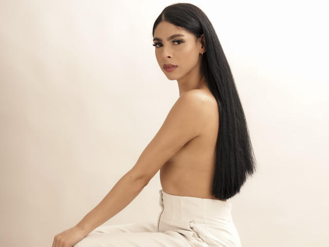 StormeAss, con lunghi capelli neri, senza top, indossa pantaloni bianchi, guarda oltre la spalla, mostrando il seno, con un'espressione neutra, in un ambiente minimalista..