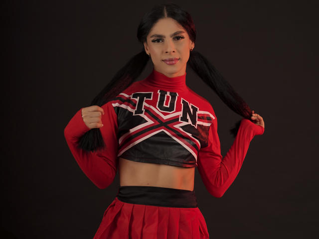 StormeAss con le trecce, indossa una maglietta e una gonna da cheerleader rosse, tiene la maglietta sollevata per mostrare la parte inferiore del ventre, rivelando i suoi addominali scolpiti, con un sorriso sicuro su uno sfondo scuro..