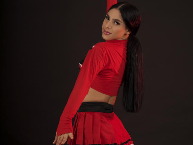 StormeAss con lunghi capelli neri, top rosso e gonna rossa, che guarda indietro, mani sui glutei, sorridente, sfondo scuro, illuminazione rossa, watermark Brazzers..