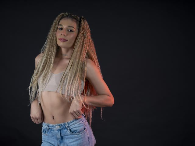 ElyassDick met lange blonde vlechten, kleine borsten in een beige topje en blauwe jeans, staand voor een donkere achtergrond, met een blik iets opzij..