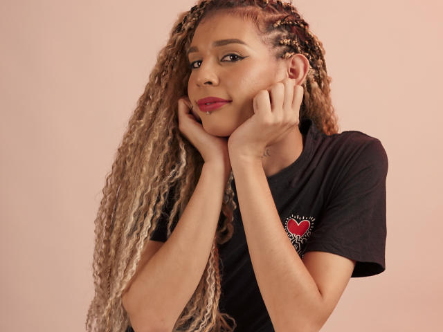 ElyassDick met lange blonde vlechten, een zwart T-shirt, handen onder de kin, roze lippenstift, een subtiele glimlach, zachte bruine huid en een schattig gezicht..