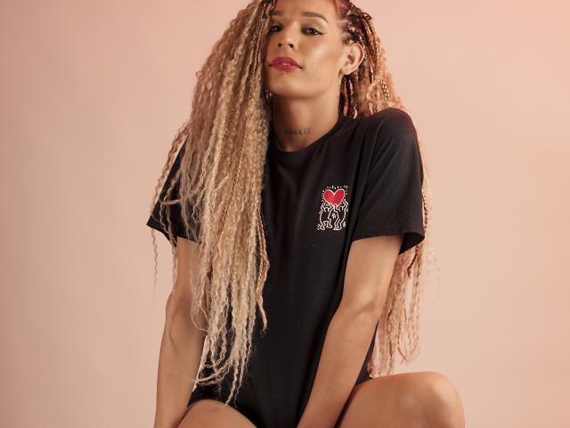 ElyassDick, com longas tranças loiras, vestindo uma camiseta preta, sentada com as pernas afastadas, mostrando um pequeno coração vermelho e um emblema de gato branco na camiseta, lábios rosados..