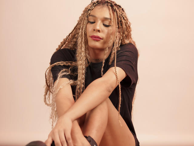 ElyassDick with long blonde braids, wearing a black shirt, sitting with knees up, looking down, has red lipstick, and a tattoo on her neck, in a soft pink background.