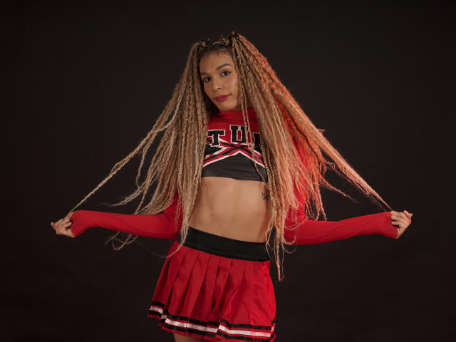 ElyassDick met lange, blonde, gevlochten vlechtjes, gekleed in een rood en zwart cheerleaderoutfit, waarbij ze de zijkanten van haar outfit vasthoudt, waardoor haar buik zichtbaar is, op een zwarte achtergrond..