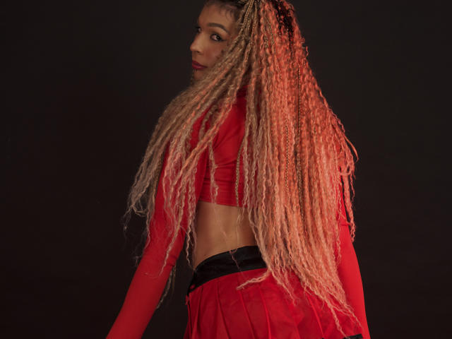 ElyassDick com longas tranças loiras e cacheadas, blusa cropped vermelha de manga comprida e saia vermelha, olhando por cima do ombro, fundo escuro, iluminação vermelha nos cabelos..