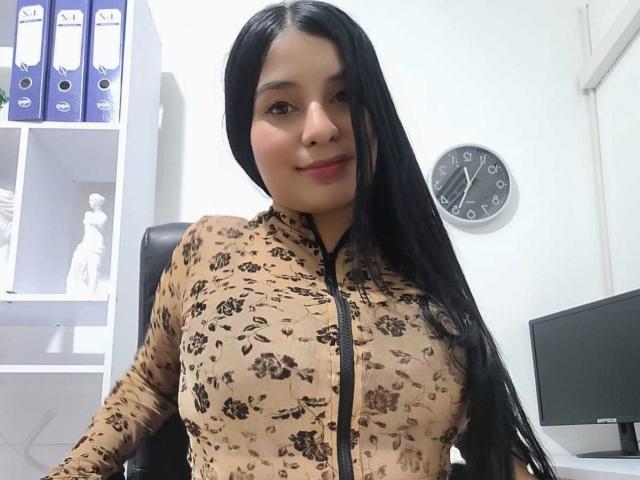 AliceNova with long black hair, smiling, wearing a tight beige floral zip-up dress, showing her big boobs, sitting in an office, clock on white wall behind her.