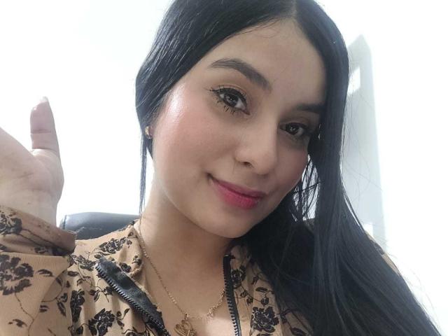 AliceNova with long black hair, brown floral dress, pink lipstick, and winged eyeliner, giving a thumbs-up, her cute face showing a slight smile, and her tits barely visible.