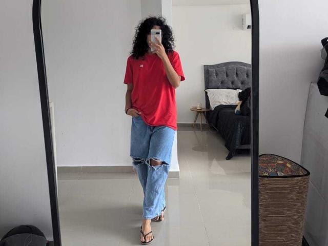 VivienneLaurent with curly black hair, wearing a red t-shirt and ripped blue jeans, taking a mirror selfie, standing in a room with a grey tufted bed and a rug.