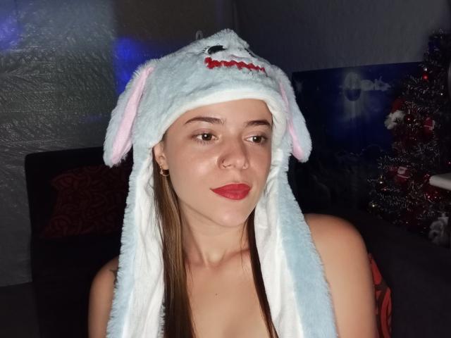 ZoeBlitz, con la pelle chiara, gli occhi marroni e il rossetto rosso, indossa un cappello bianco a forma di cane soffice con orecchie rosa e naso rosso; le sue spalle e il petto sono scoperti..