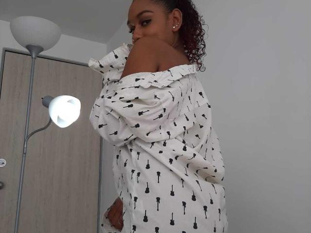 CandyEsme, met donkere huid, krullend rood haar, een witte blouse met een open schouder en zwarte kwasten, kijkt over haar schouder en staat voor een lamp en een deur..