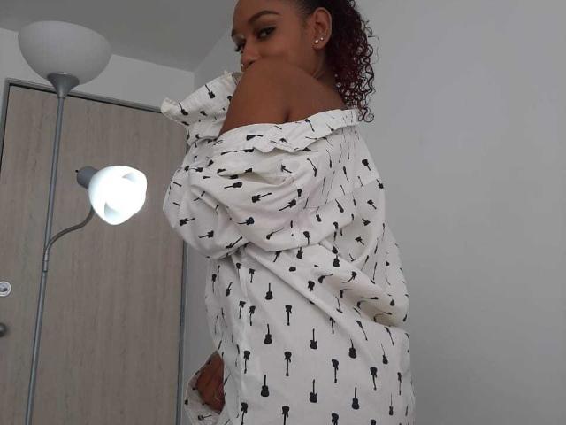 CandyEsme, com pele escura, cabelo ruivo encaracolado, camisa branca com decote ombro a ombro com estampas de guitarras pretas, olhando por cima do ombro, em pé em frente a um abajur e uma porta..