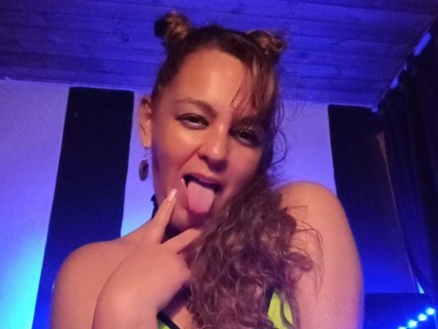 AmandaHotL with double buns, sticking out her tongue, and touching it with her finger, showing off her cute face and curly brown hair. She's wearing a black top.