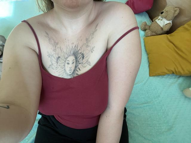 DiLaria's tits, covered in a sun face tattoo with rays, are visible above a red tank top, and she's wearing black pants, with a teddy bear in the background.