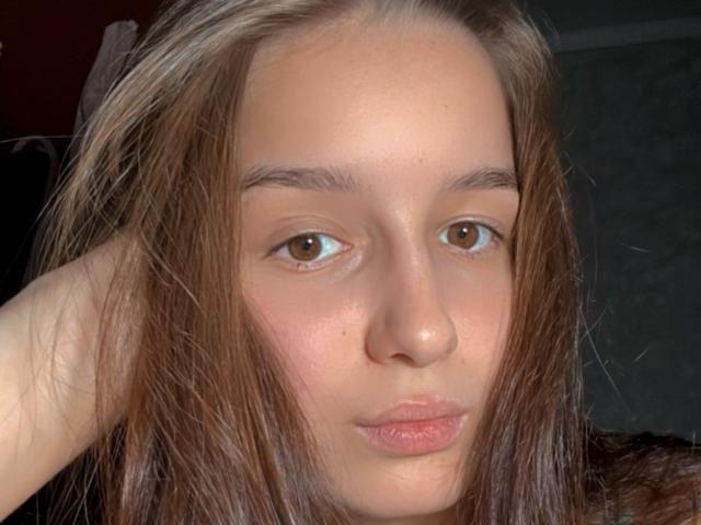 Het gezicht van TeresaCatty, met bruine ogen en lichtjes opgetrokken, roze lippen, lang bruin haar, met een hand in haar haar, en een lichte huid, die recht in de camera kijkt..