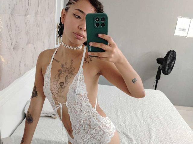 SilvanaXOwen wearing a white lace lingerie bodysuit, showing tattoos, small tits, and a choker, taking a mirror selfie with a green phone, standing in a bedroom.