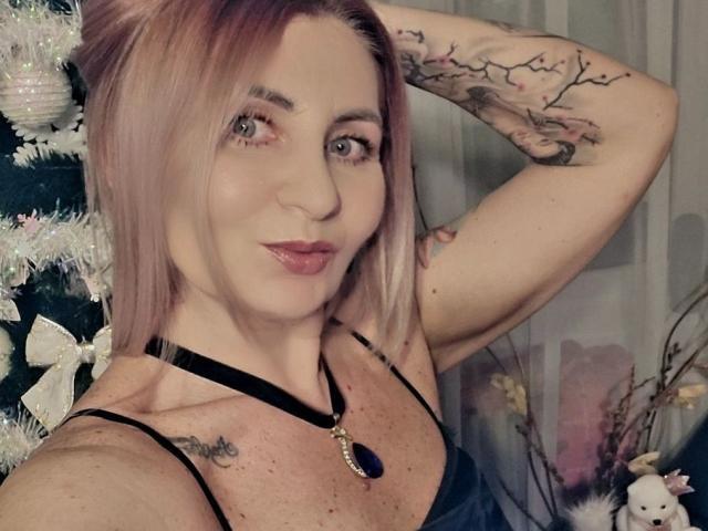 ChaudeEvely with pink hair, tattoos on arms, black choker with purple gem, black spaghetti straps, smiling, hand behind head, showing small tits and freckles.