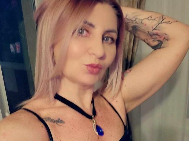 ChaudeEvely with pink hair, pouty lips, black strap top, blue gem necklace, arm raised showing tattoo, green eyes, slight freckles, looking at camera, in dim lighting.