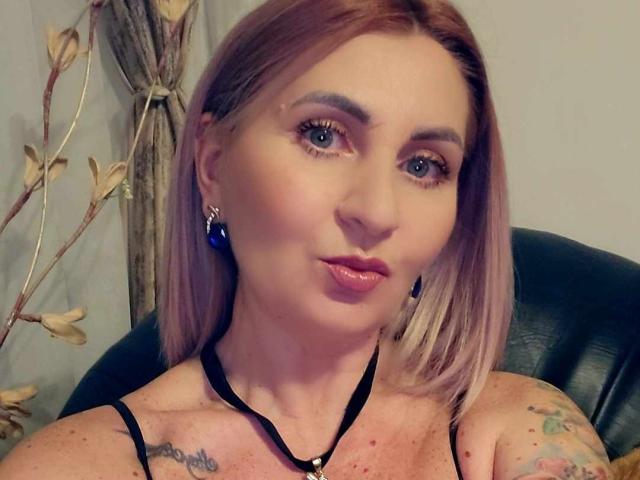 ChaudeEvely has blue eyes, pink lipstick, and a black halter top, with blonde bob hair, wearing blue earrings, showing tattoos on her shoulders, pouty face.