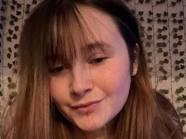 MaggieMoll with straight brown hair, nose ring, and lower lip piercing, smiling softly, has a slight blush and a relaxed, cute, and sexy appearance.