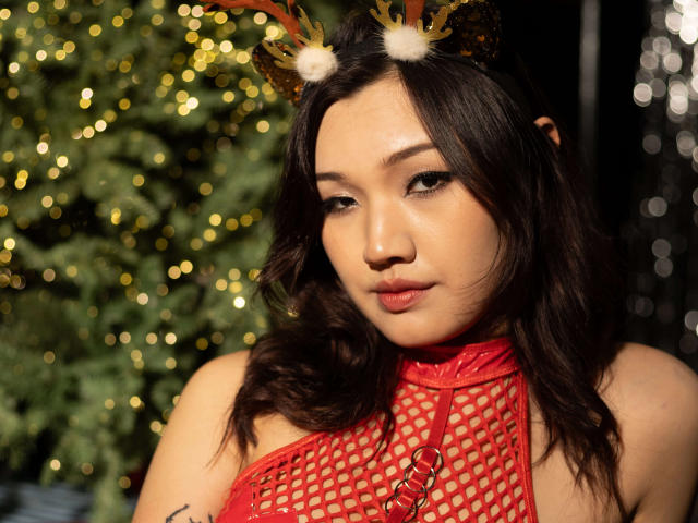 ShadowStrike has a cute face, dark wavy hair, and wears a red fishnet top with reindeer antlers. Her pouty lips and shiny skin are sexy. Boob outline visible.