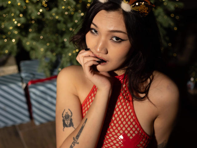 ShadowStrike with dark hair, red fishnet top, and reindeer headband, biting her finger, showing tattoos on her arm, looking seductively at the camera. Her tits are visible.