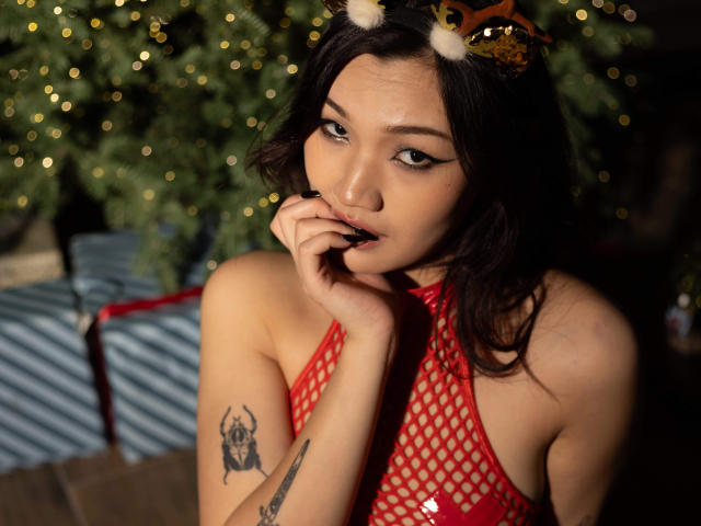ShadowStrike with cat ears, red fishnet top, biting finger, tattoos on left arm, looking seductively at camera, dark wavy hair, slightly open mouth, Christmas tree blurred in background.