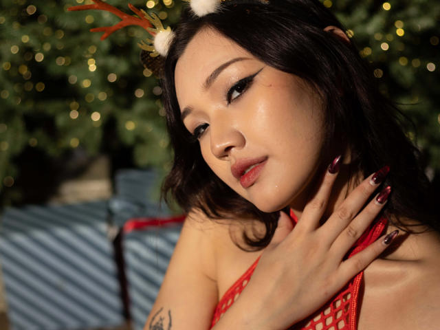 ShadowStrike with dark hair, red lipstick, and red fishnet top, wearing reindeer antlers, touching her neck, showing tattoos, with glossy skin, and dark red nails, looking seductively.