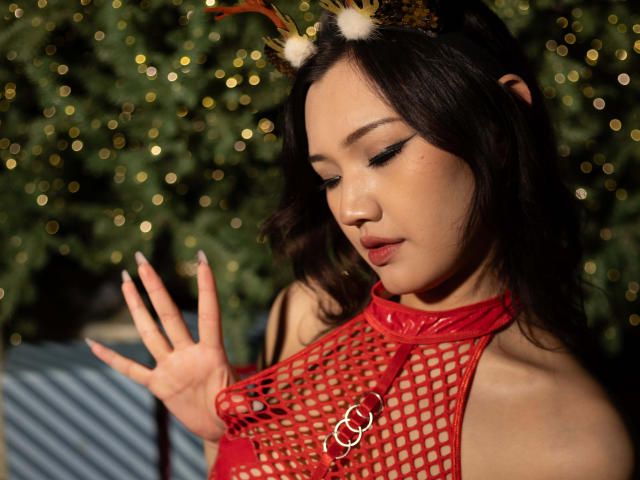 ShadowStrike with dark hair, wearing red fishnet top, antler headband, and green eyeshadow, touching her chest, showing small, perky tits, and a cute, sexy face, in a Christmas setting.