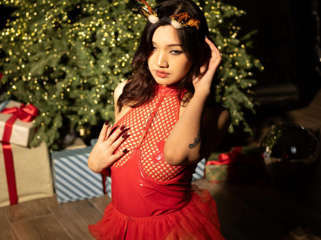 ShadowStrike with wavy black hair, red fishnet dress, red tulle skirt, and reindeer headband, touching her tits, showing a tattoo on her right arm, in warm sunlight.