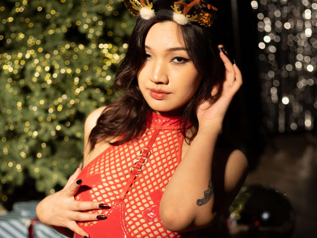 ShadowStrike with dark wavy hair, wearing a red fishnet top, reindeer antlers, and black nails, holding her tits, tattoo on her right arm, looking seductive in a warm light.