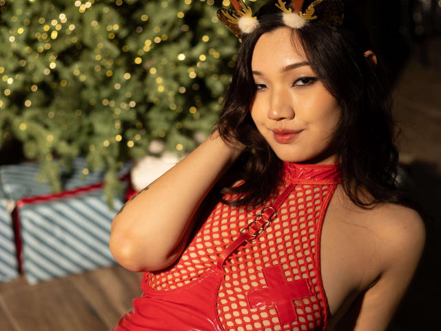 ShadowStrike with dark hair, red fishnet dress, and reindeer antlers, showing a sly smile, has small tits and a cute face, standing in front of a Christmas tree.