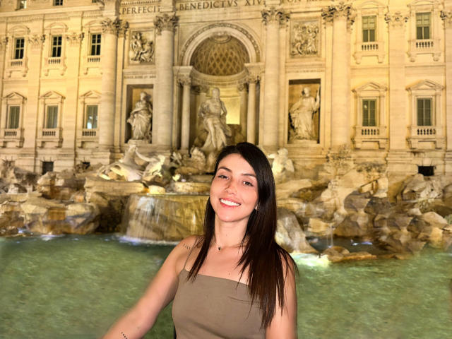 AlanaBazan, with dark brown hair and smiling, wears a strapless brown dress, standing in front of a detailed stone fountain, water visible in the foreground.