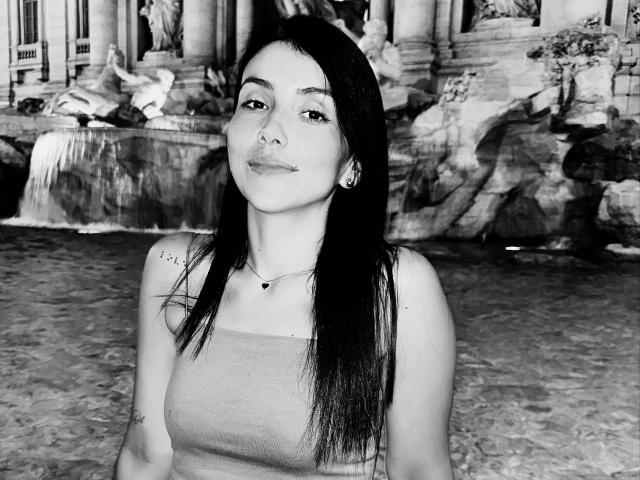 Black-and-white photo of AlanaBazan with long dark hair, small tits, wearing a tank top, standing with a confident smile, small nose ring, and a necklace.
