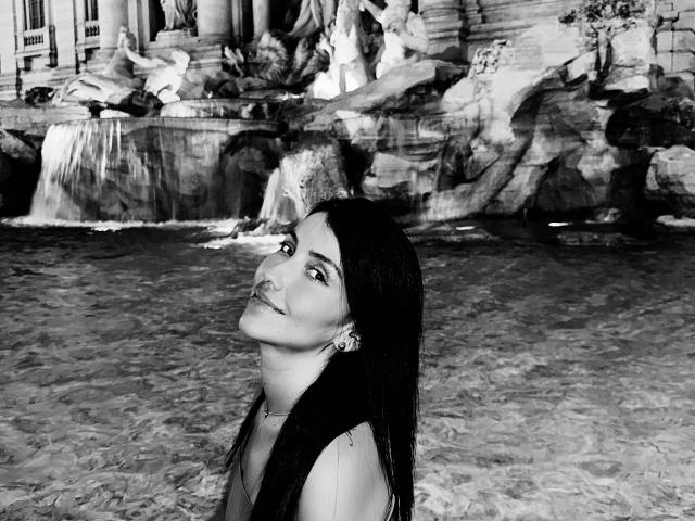 Black and white photo of AlanaBazan with long dark hair, looking up with a slight smile, showing her neck and earrings, standing in shallow water. Boobs not visible.