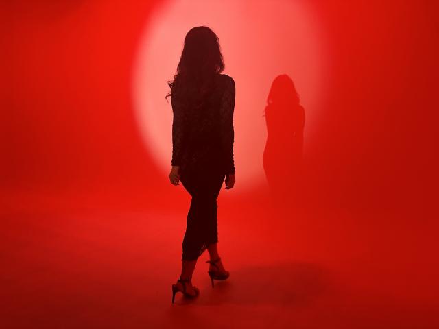 Silhouette of AlanaBazan in black high heels and tight pants, standing with back to camera, red light casting shadow on wall, wavy dark hair, confident stance.