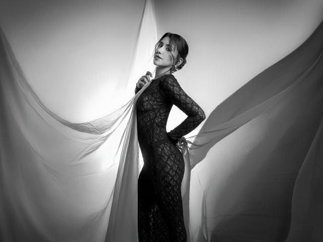 Black-and-white photo of AlanaBazan in a lace, long-sleeve, form-fitting dress, holding a sheer fabric, standing with hand on hip, looking up, with a soft light behind.