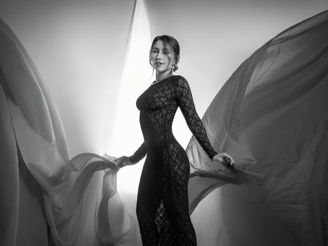 AlanaBazan with big tits, wearing a black lace dress, holding sheer fabric, standing in front of a bright light, smiling, black-and-white photo.