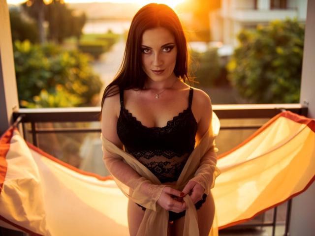 AmandaChilli with dark brown hair, black lace bra, and panties, holding a beige robe, standing on a balcony at sunset, sunlight in the background. Boobs, lace, and robe.