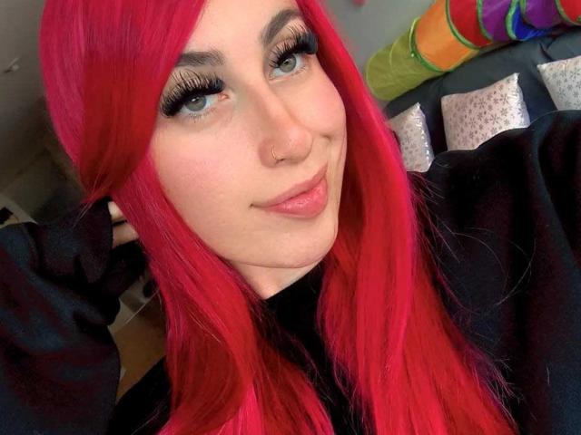 SammyViolet with bright red hair, blue eyes, and long black eyelashes, wearing a black shirt, nose ring, and a slight smile, showing a cute, sexy face.