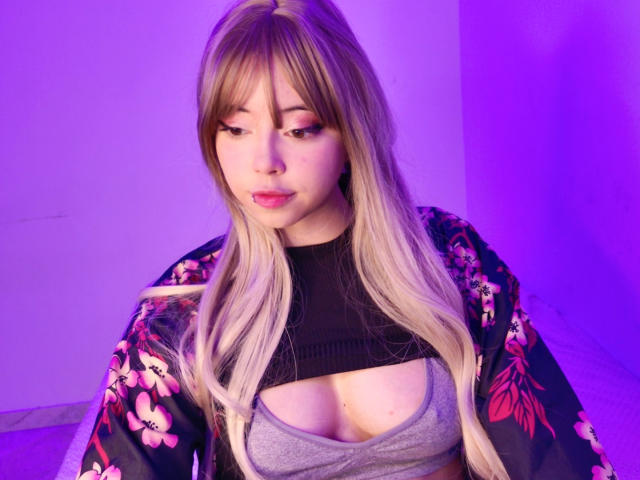 Arieyaara mit langen blonden Haaren, die einen schwarzen Kimono mit Blumenmuster trägt, ein graues Oberteil mit Ausschnitten, das ihre Brüste zeigt, violette Beleuchtung, die nach unten schaut mit pinkem Lippenstift und einem Nasenring..